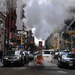 A photo showing a busy street with cars lining up either side of a pipe with plumes of steam floating above it. 