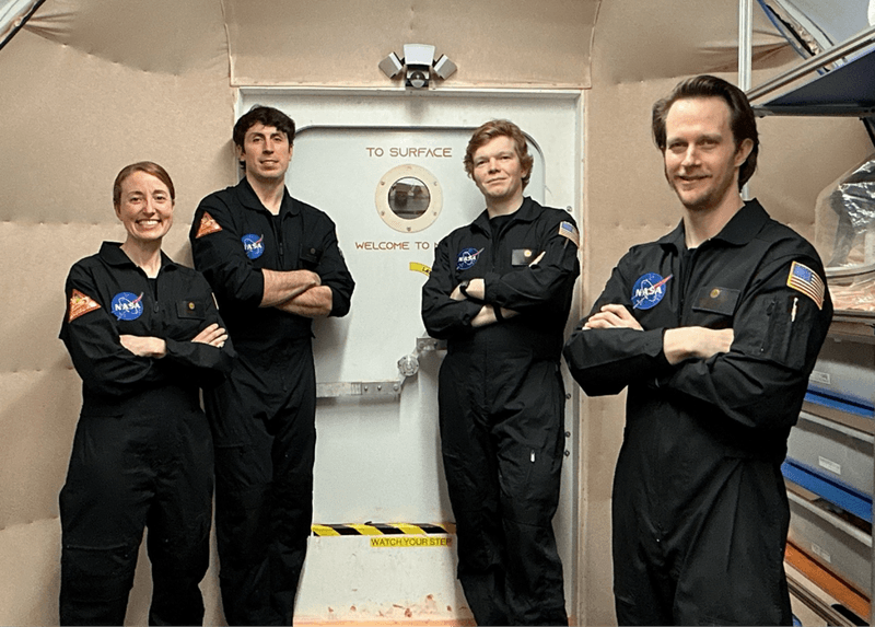 four white people, one short woman and three tall men, standing in front of a habitat door