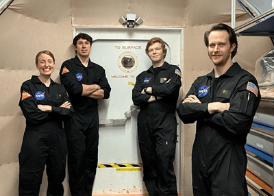 four white people, one short woman and three tall men, standing in front of a habitat door