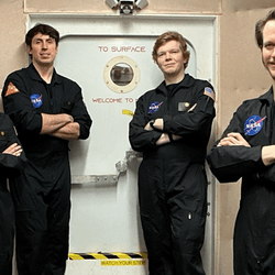 four white people, one short woman and three tall men, standing in front of a habitat door