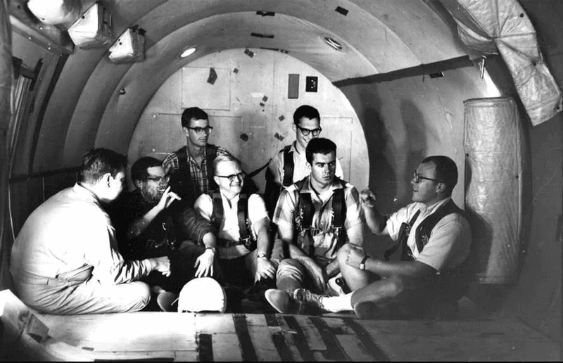 seven men sitting in an empty aircraft, with walls and floor padded.
