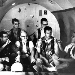 seven men sitting in an empty aircraft, with walls and floor padded.