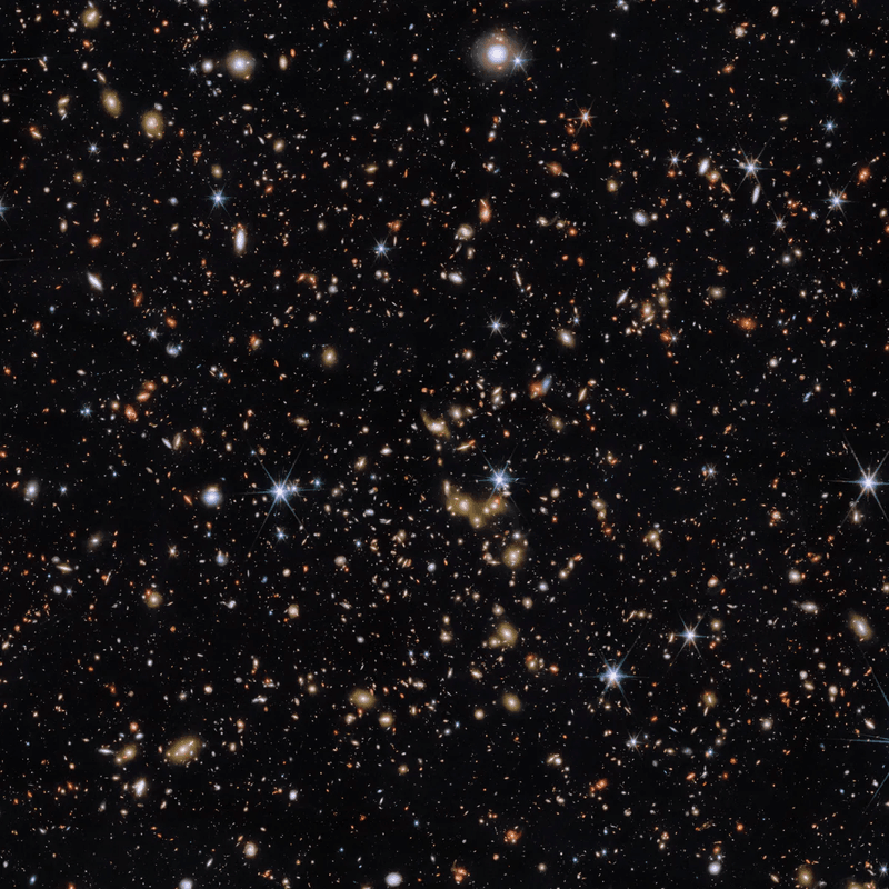 An area of deep space with thousands of galaxies in various shapes and sizes on a black background. Most are circles or ovals, with a few spirals. More distant galaxies are redder in colour and smaller, down to being mere dots, while closer galaxies are a bit larger and white or blueish. A few gold-coloured galaxies are bunched closely together in the centre. Bright stars surrounded by spikes lie in our galaxy.]