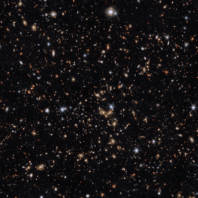 An area of deep space with thousands of galaxies in various shapes and sizes on a black background. Most are circles or ovals, with a few spirals. More distant galaxies are redder in colour and smaller, down to being mere dots, while closer galaxies are a bit larger and white or blueish. A few gold-coloured galaxies are bunched closely together in the centre. Bright stars surrounded by spikes lie in our galaxy.]