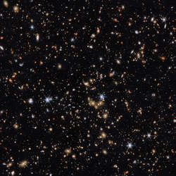 An area of deep space with thousands of galaxies in various shapes and sizes on a black background. Most are circles or ovals, with a few spirals. More distant galaxies are redder in colour and smaller, down to being mere dots, while closer galaxies are a bit larger and white or blueish. A few gold-coloured galaxies are bunched closely together in the centre. Bright stars surrounded by spikes lie in our galaxy.]